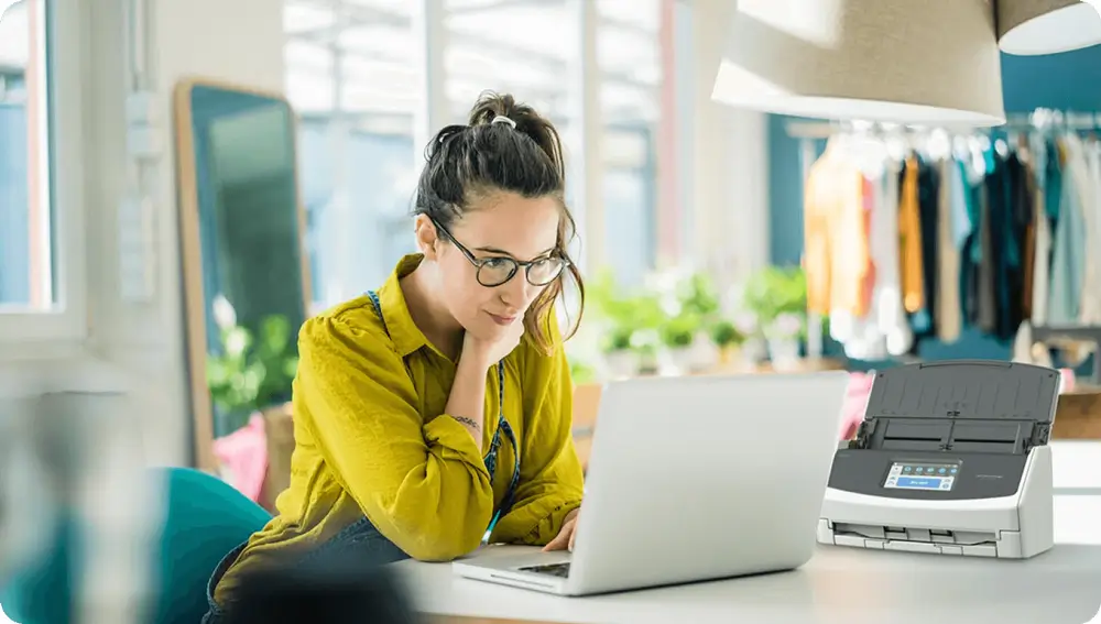 Young woman in a bright, creative workspace using a laptop with a compact document scanner on the desk beside her.